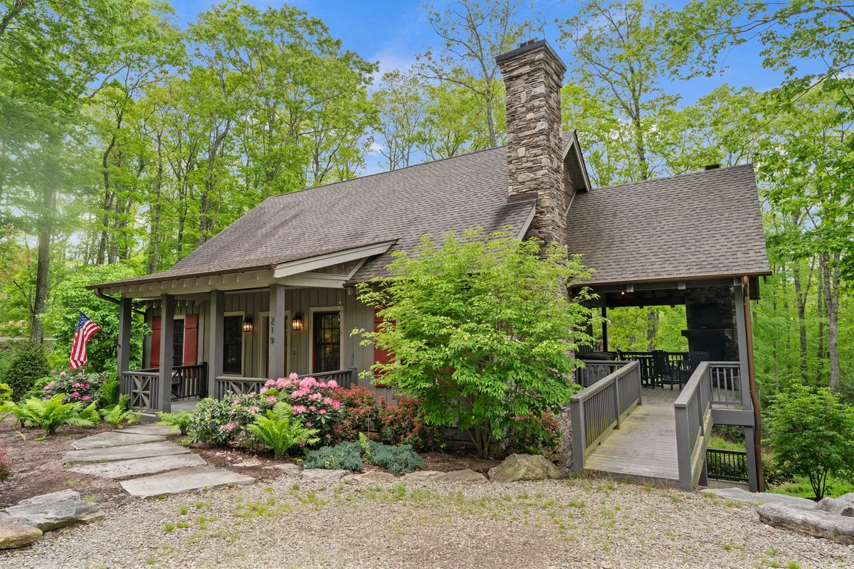 This exterior view showcases a wooden porch surrounded by lush greenery and flowering shrubs, with a stone chimney highlighting the home's rustic charm. The inviting space is ideal for family gatherings, nature walks, and peaceful evenings.