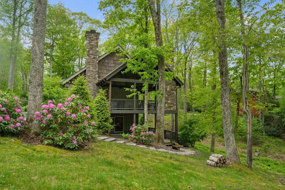 This exterior image features a charming stone chimney, a welcoming front porch, and flowering shrubs, all set against a peaceful natural backdrop. The porch offers a serene spot for outdoor relaxation, and the lush landscaping hints at the surrounding hiking trails and nature walks available for guests and their pets.