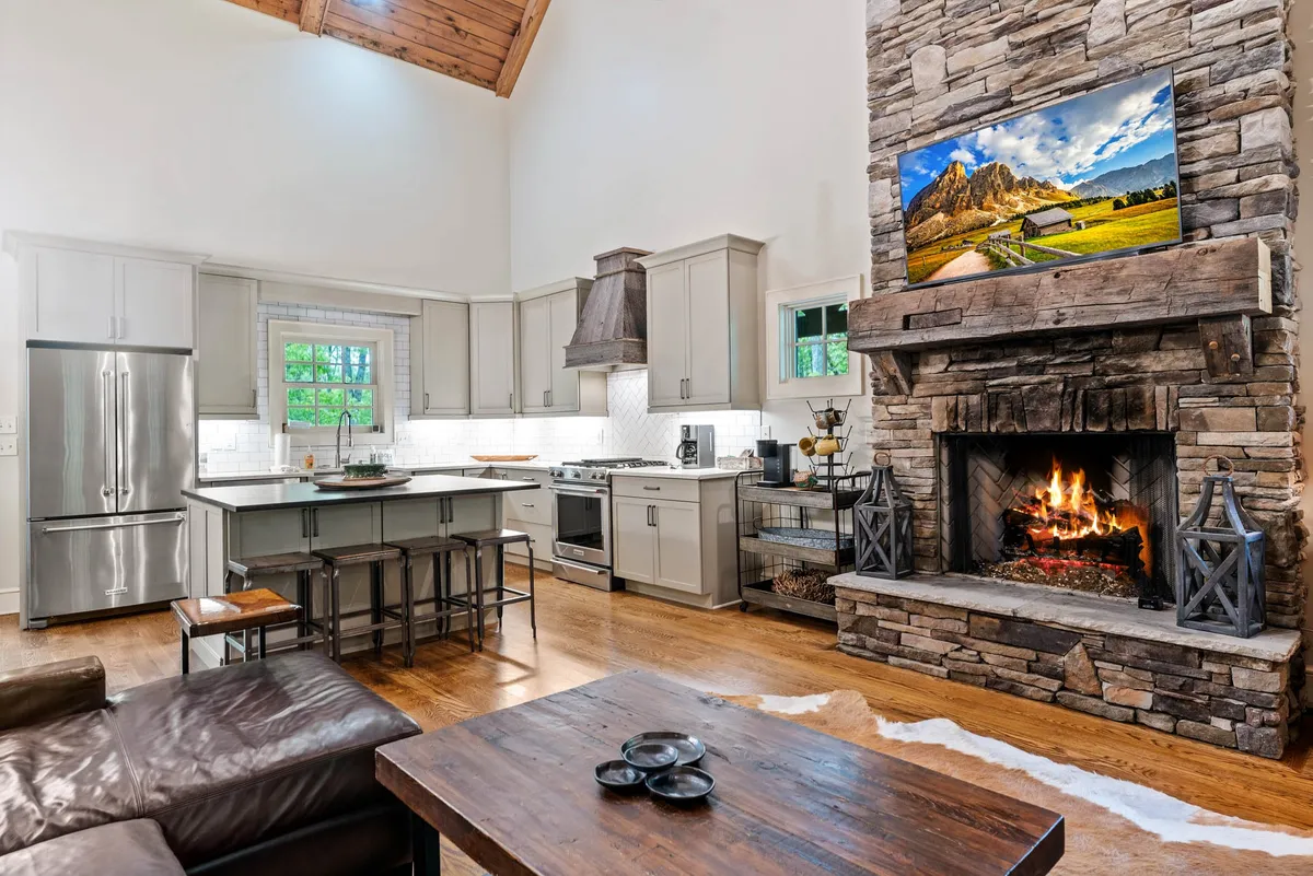 An expansive view of the open-concept living area showcasing a striking stone fireplace, a spacious kitchen island with bar seating, and a large rustic wooden dining table. The scene is anchored by inviting lighting and the warmth of a flickering fire, creating a welcoming atmosphere for gatherings.