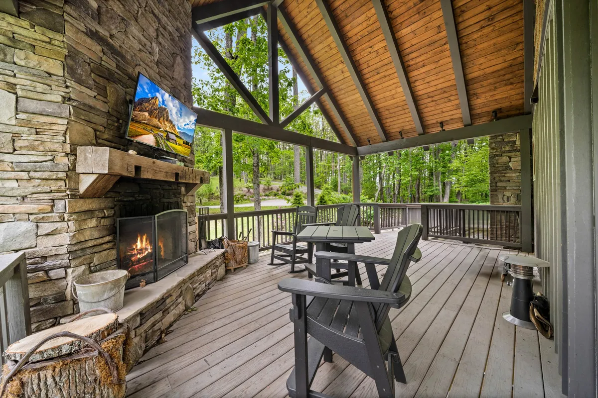 A spacious wooden deck featuring a beautifully crafted stone fireplace, surrounded by comfortable outdoor furniture and a television mounted above the mantle. The area is framed by lush greenery, inviting guests to relax by the fire and enjoy the outdoors in any season.