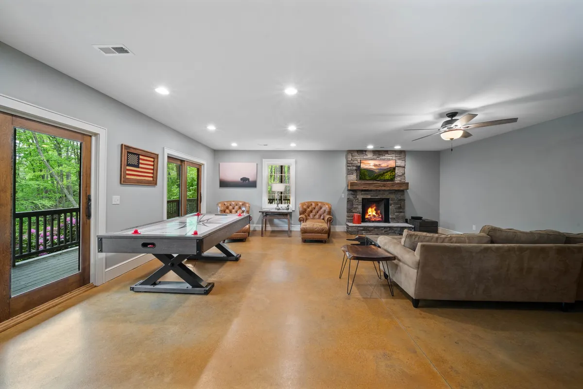 This image features a lively game room with an air hockey table, a cozy fireplace, comfortable seating, and large windows letting in natural light. The arrangement invites both play and relaxation, with elements that support friendly competition and social gatherings.