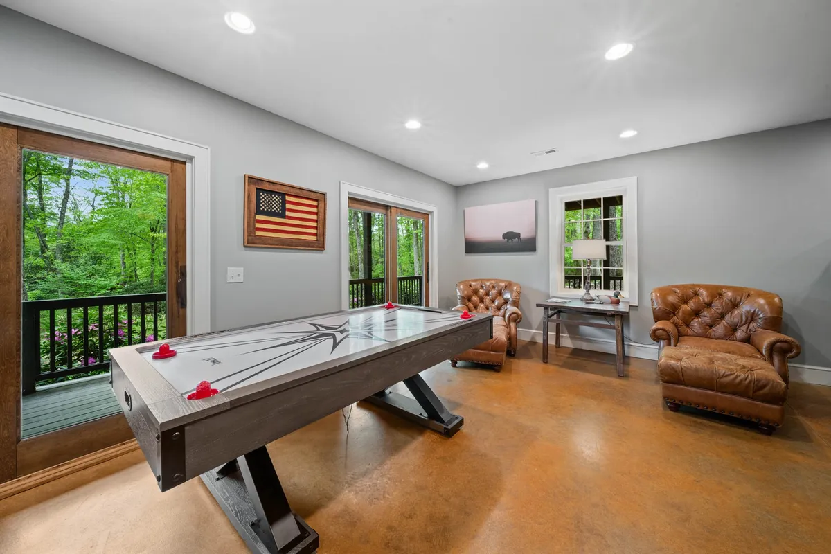 A playful and comfortable game space with an air hockey table, leather armchairs, artwork, and large windows offering serene nature views. The room is bright, airy, and designed for interactive fun and relaxation.
