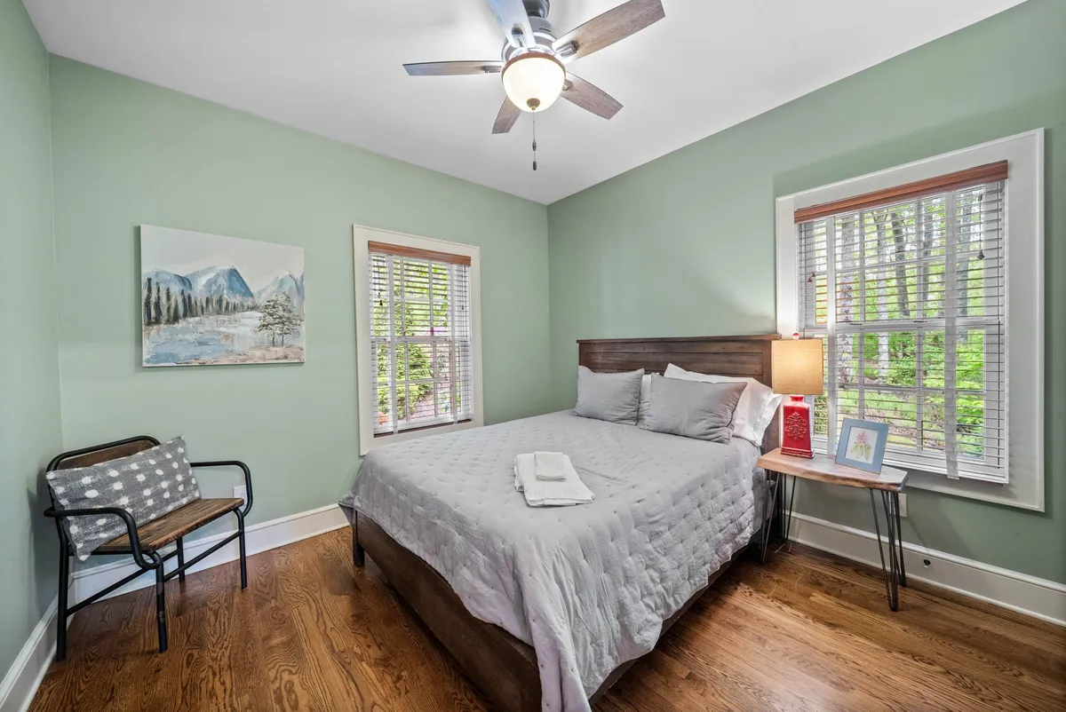 A neatly made bed with an array of pillows sits beneath artwork and windows that let in natural light. A nightstand with a lamp and book adds a cozy, lived-in feel, while the serene decor fosters rest and relaxation.