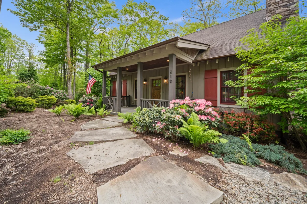 The charming front porch of a home, bordered by flower beds and featuring a stone pathway and American flag. The setting invites relaxation, with ample space for family gatherings or simply enjoying the surrounding nature.