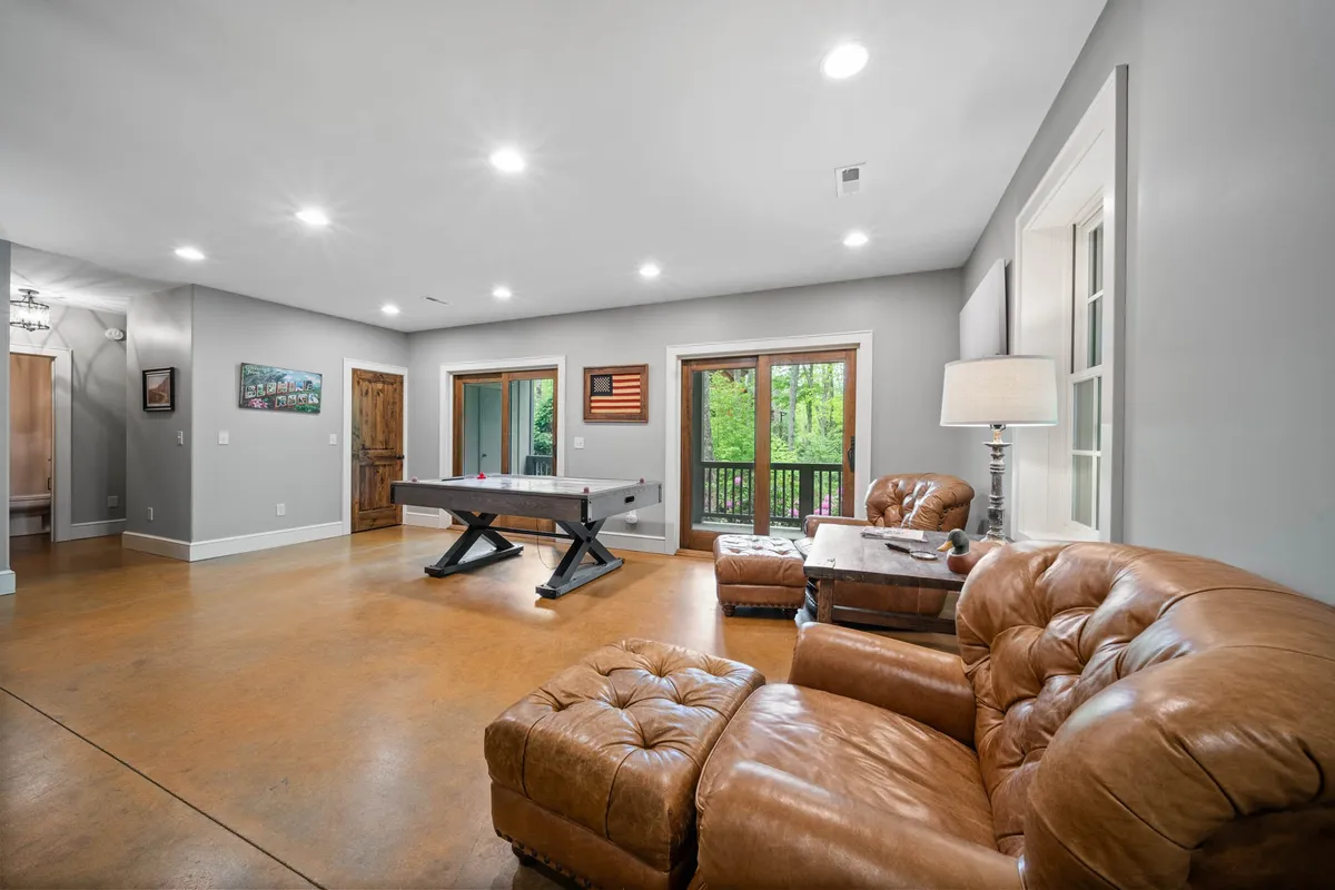 A spacious game room interior with a pool table, leather couch, large windows that let in plenty of natural light, and a table lamp. The room is set for fun activities like playing pool or gathering for movie nights, with a cozy ambiance ideal for family bonding.