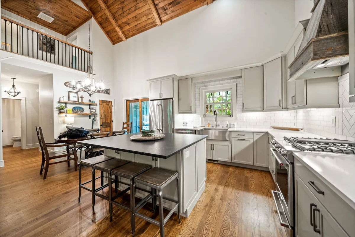 A cozy perspective of the kitchen and dining area, featuring a prominent kitchen island, exposed wooden beams, a large wooden dining table, and sunlit windows that fill the space with natural light. The room exudes warmth and invites togetherness.