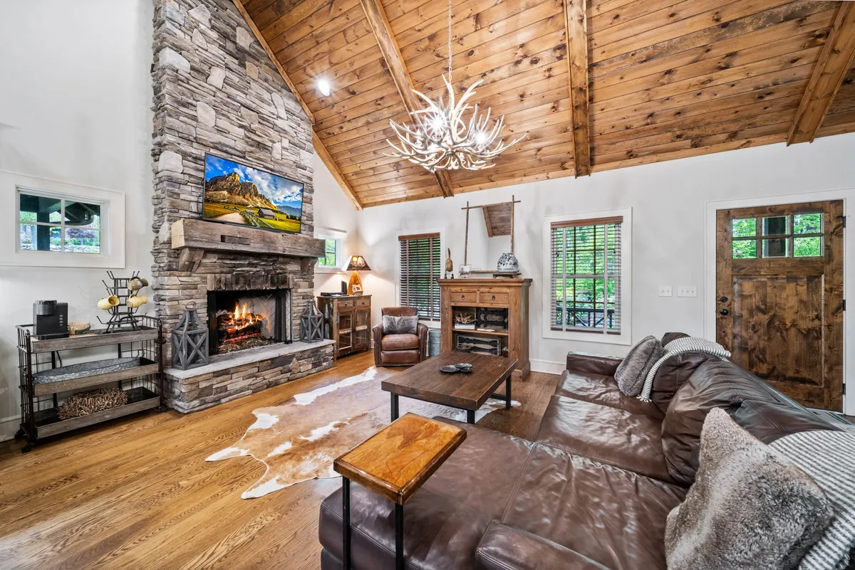 The image showcases a warm and inviting living room with a striking floor-to-ceiling stone fireplace, soaring wooden ceilings, a plush leather sofa, a stylish cowhide rug, and a large flat-screen TV. Soft evening light and cozy throws invite guests to relax and unwind. The space exudes rustic elegance and comfort, perfect for gathering with family or friends.