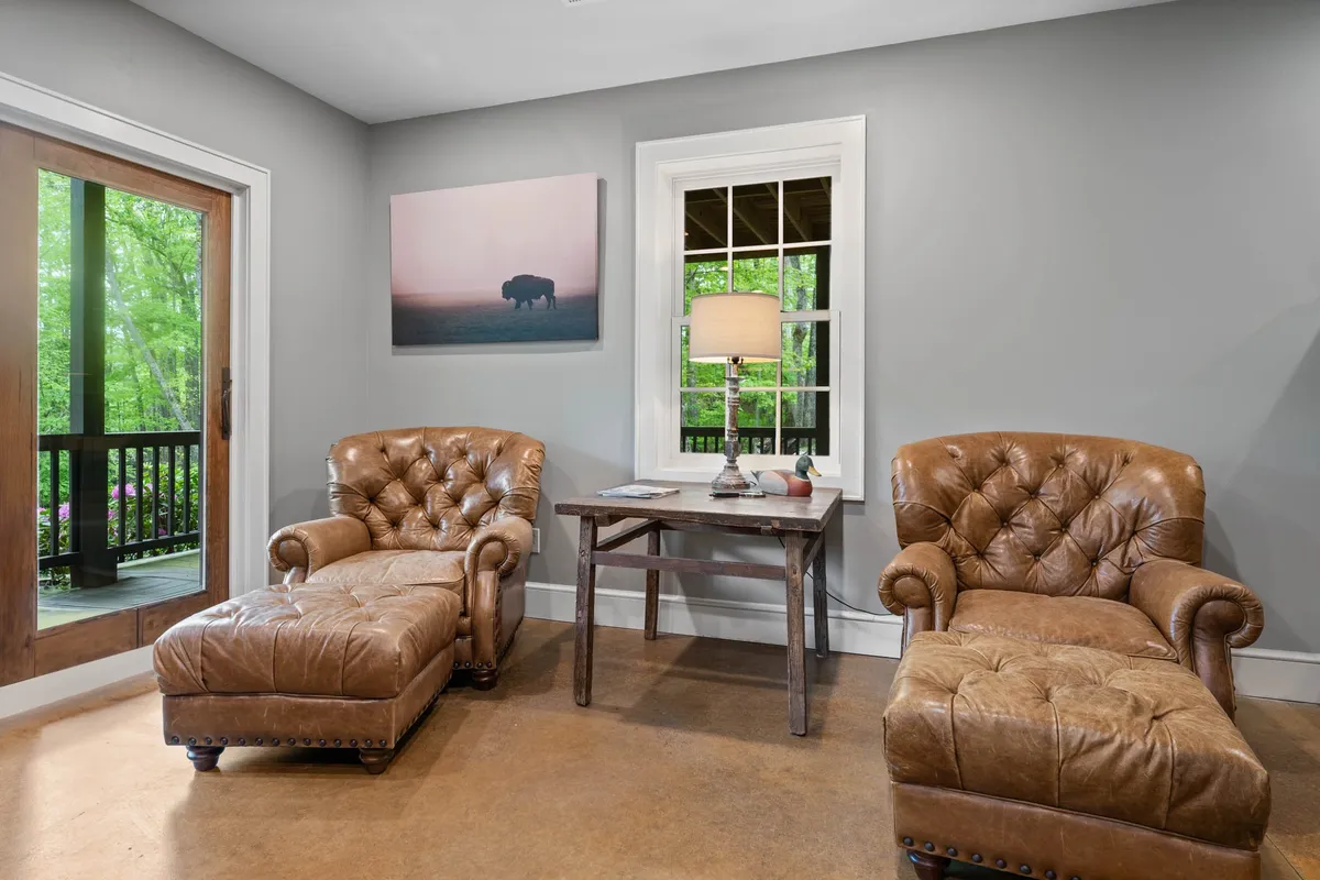 This image features a pair of comfortable leather chairs beside a wooden table and lamp, with artwork on the wall and a visible view of the outdoors through a glass door. The setting suggests a cozy spot for relaxing, socializing, and enjoying the natural beauty just beyond the threshold, blending indoor comfort with access to the outside world.