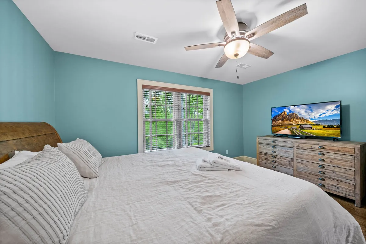 This image features a spacious king-sized bed with textured bedding, a wooden dresser, a large window with blinds, and a flat screen TV. The room is filled with soft, natural light, creating a tranquil and cozy atmosphere. The furnishings are simple yet elegant, emphasizing comfort and relaxation.